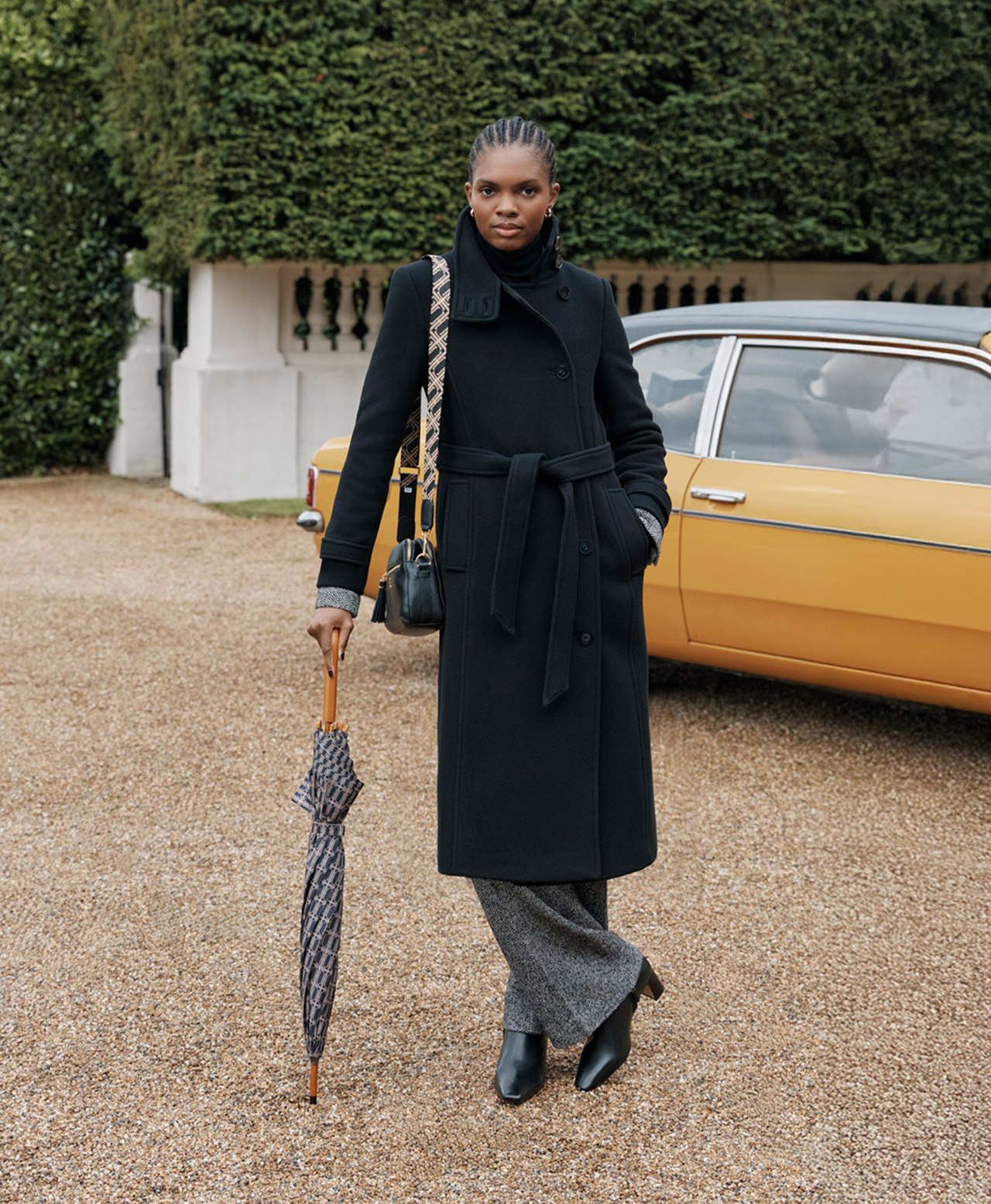  A woman wearing a long, black Hobbs coat, with a monogram umbrella and matching bag.