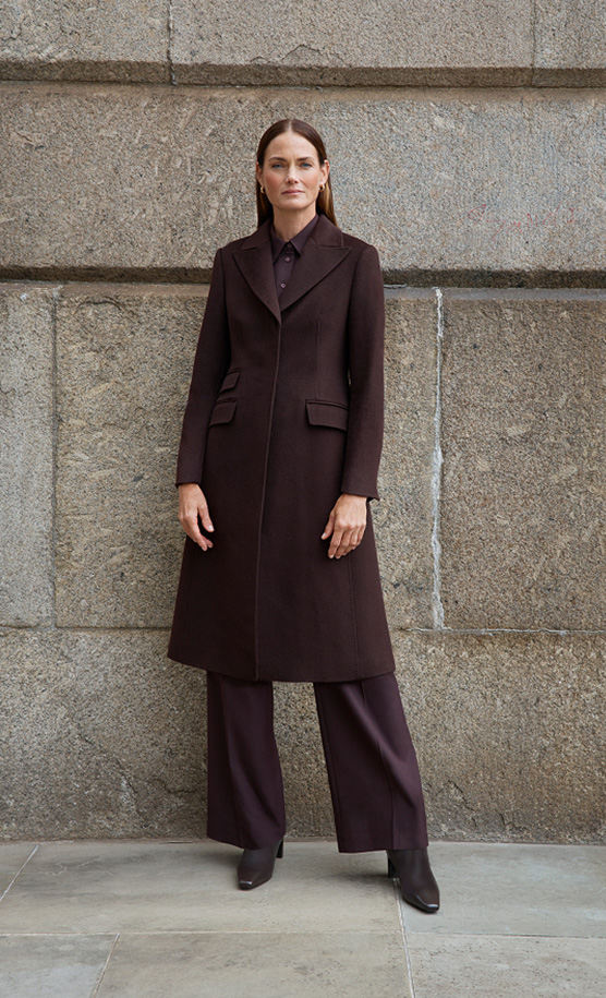 Model stands against a wall wearing a brown wool coat.