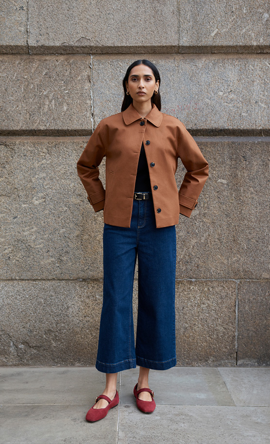 Model stands against a wall wearing a tan cropped trench.