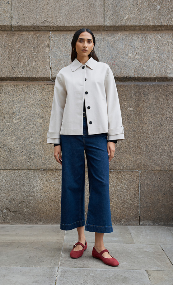 Model stands against a wall wearing a grey cropped trench.
