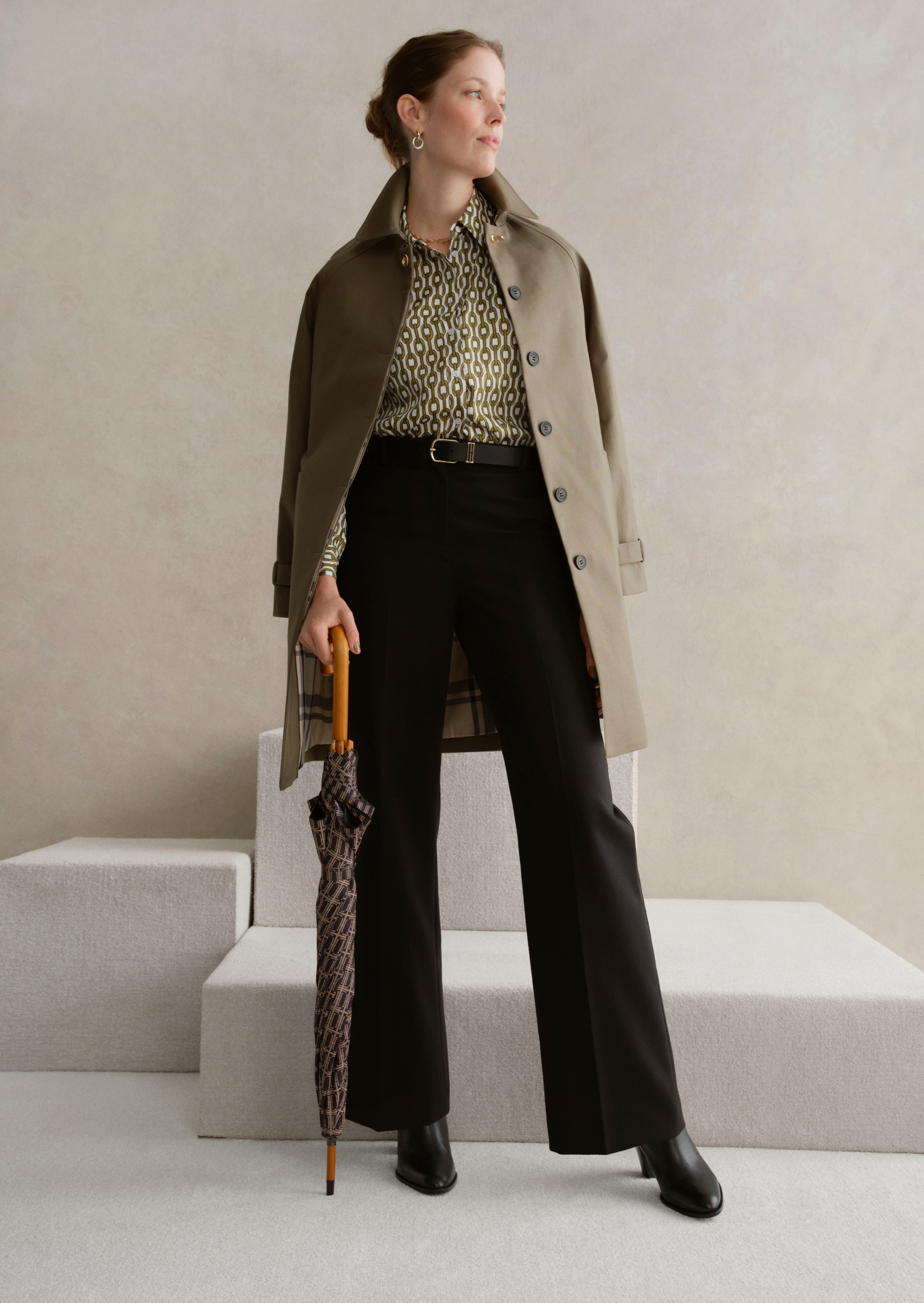 A gallery of two images displaying a model wearing a khaki trench coat, printed blouse and tailored trousers.
