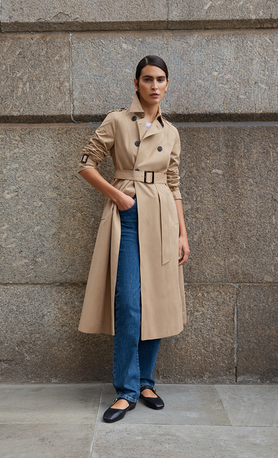 Model stands against a wall wearing a beige trench coat.