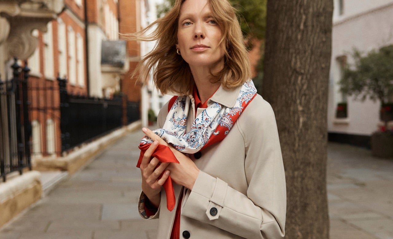 A woman wearing a beige trench and paisley red neck scarf.