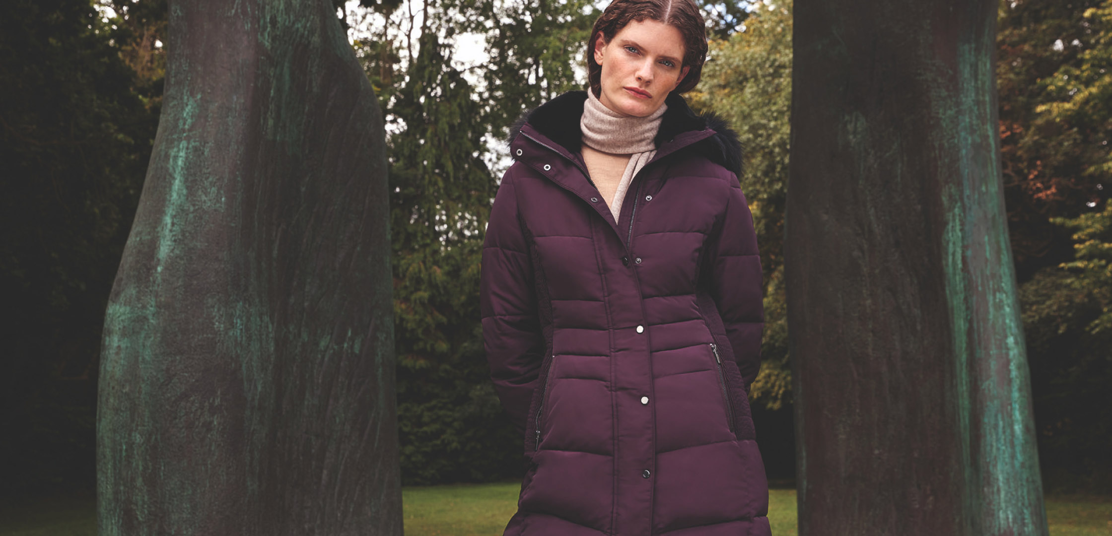 Close-up of model photographed in front of an outdoor sculpture wearing a burgundy puffer coat.
