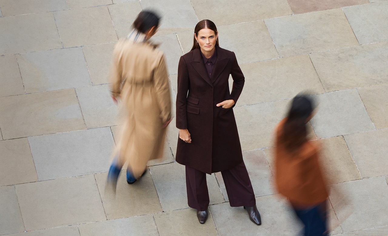 One model stands still on a city street as two other models walk past, all wearing Hobbs coats.