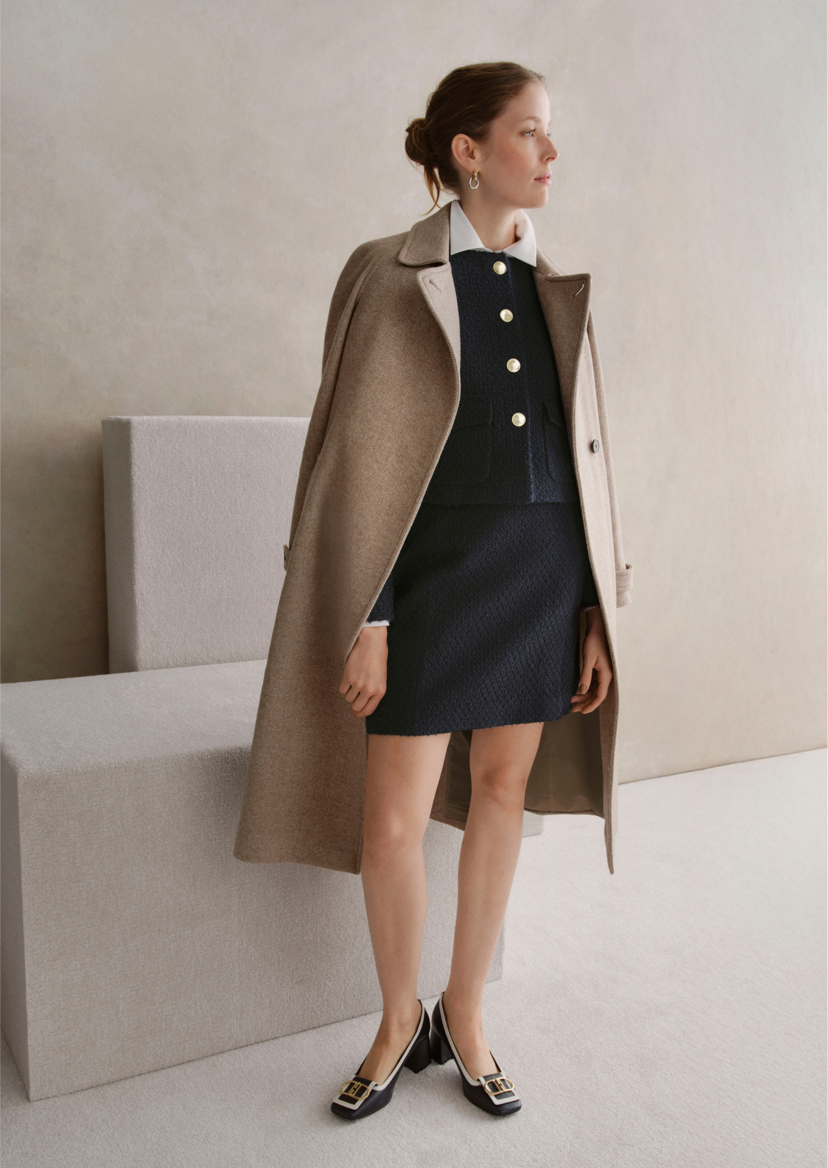 An image of a model wearing a beige tailored coat over a navy skirt and jacket