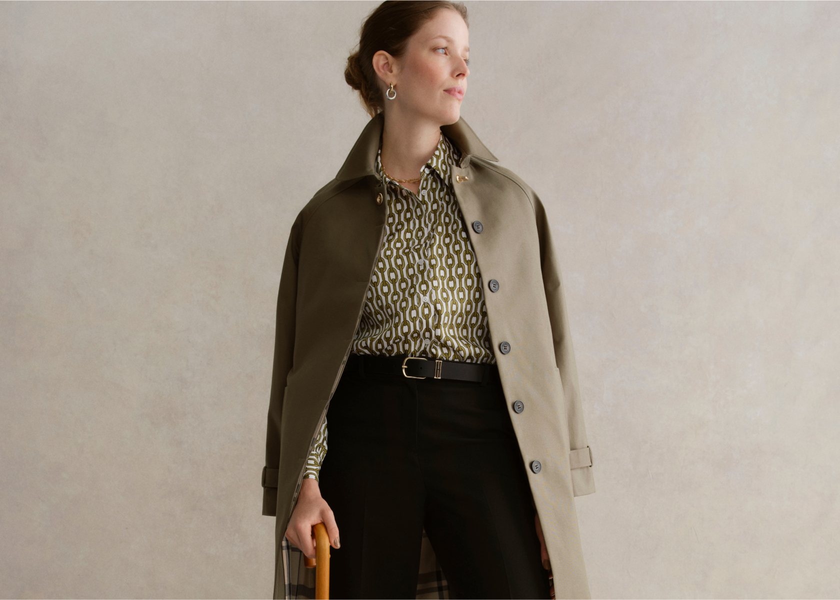 An image of a model wearing a khaki trench coat, printed blouse and tailored trousers.