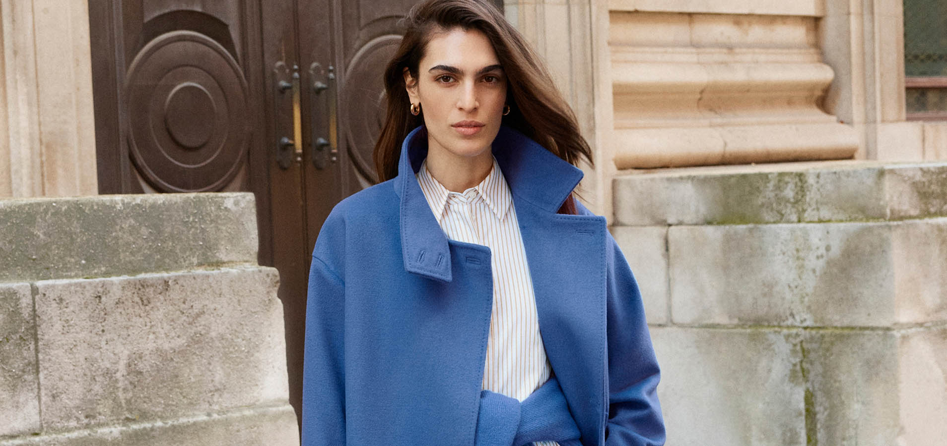 A woman wearing a blue jumper and coat, layered over brown trousers.