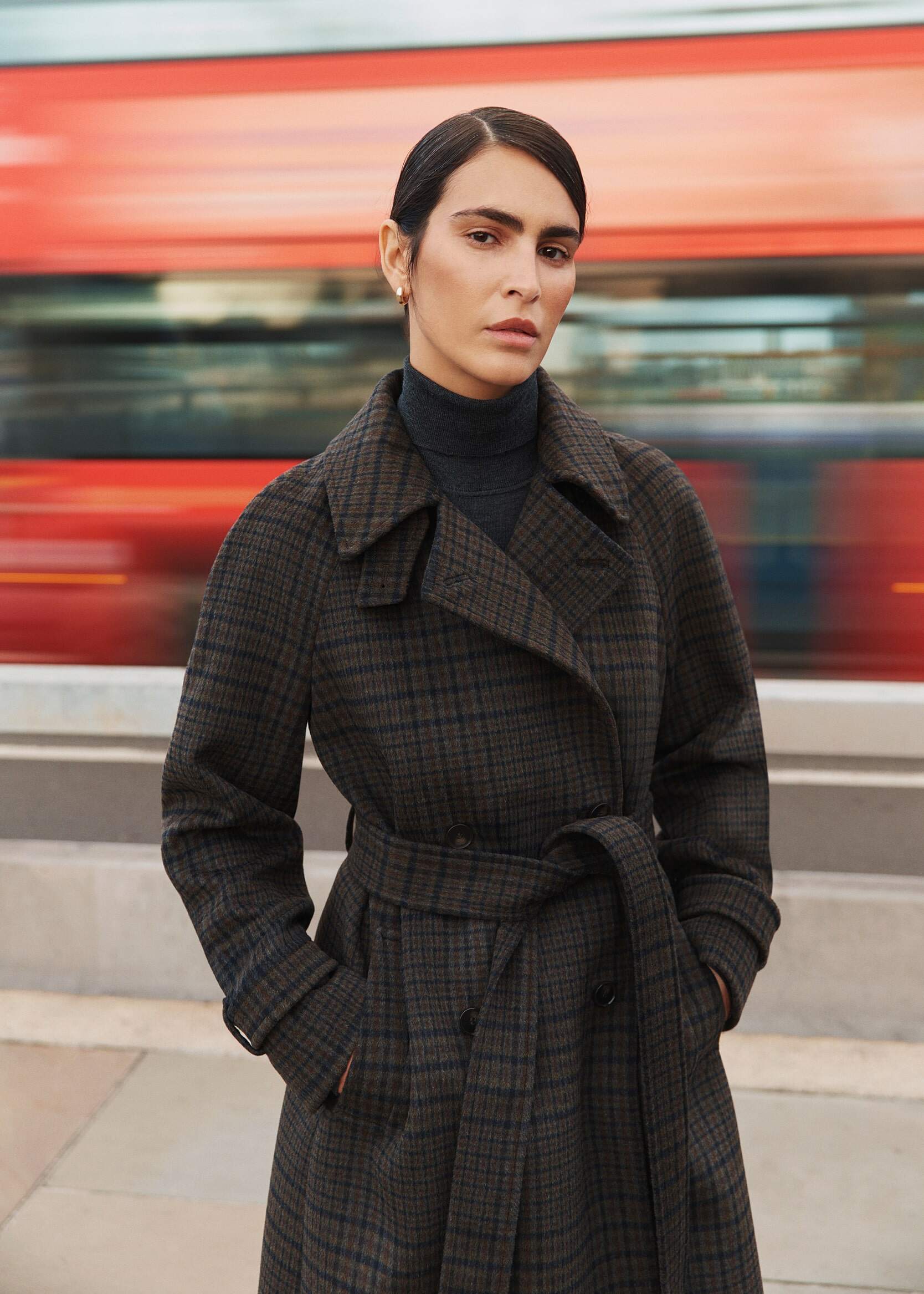 A model in a check wool coat stands still on a street, as a London bus passes by behind.