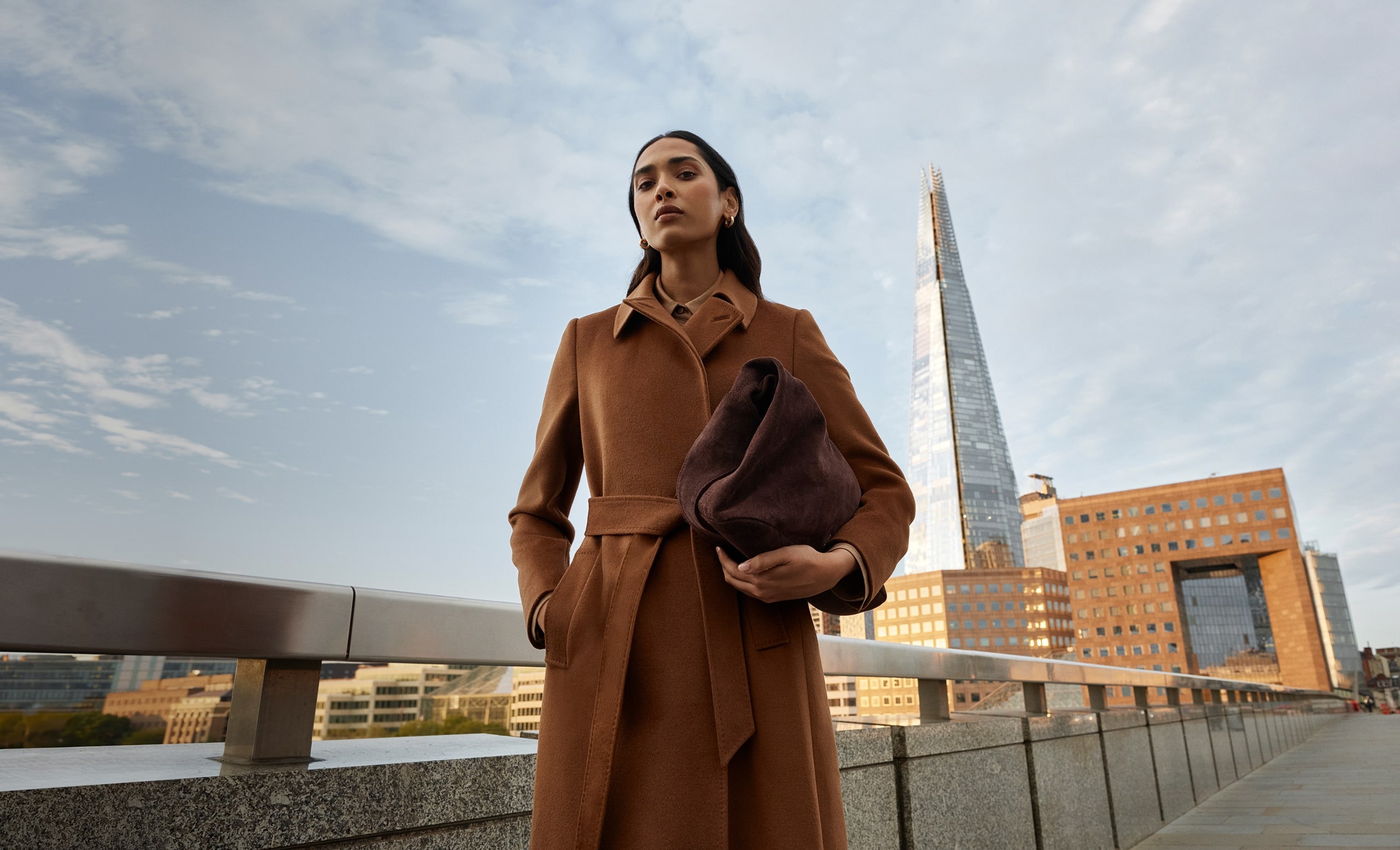 A model stands on London Bridge wearing a tan brown wrap coat.
