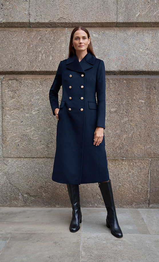 Model stands against a wall wearing a navy military coat.