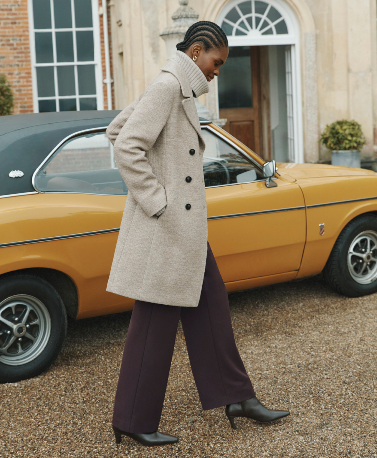 A woman wearing a beige coat layered over a high neck jumper and brown trousers.