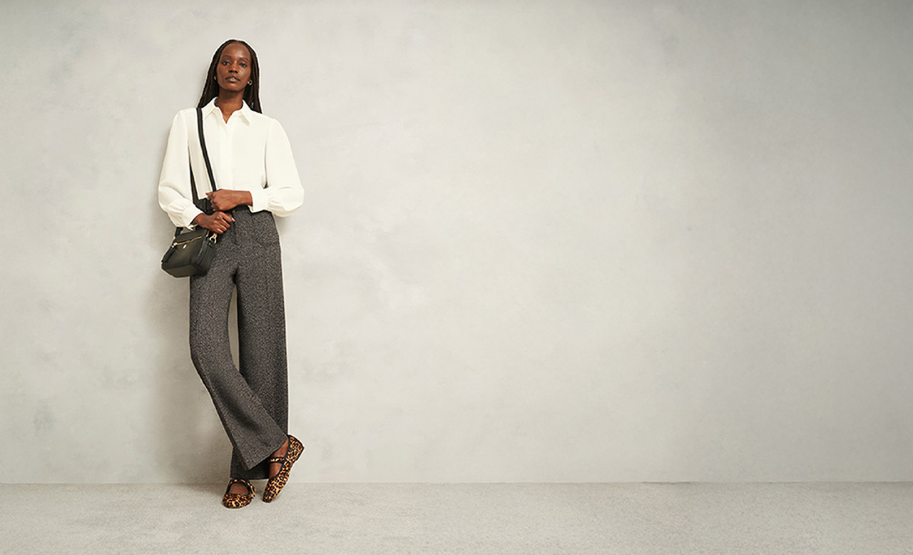 A woman wearing Hobbs grey tailored trousers, white blouse and leopard ballet flats.